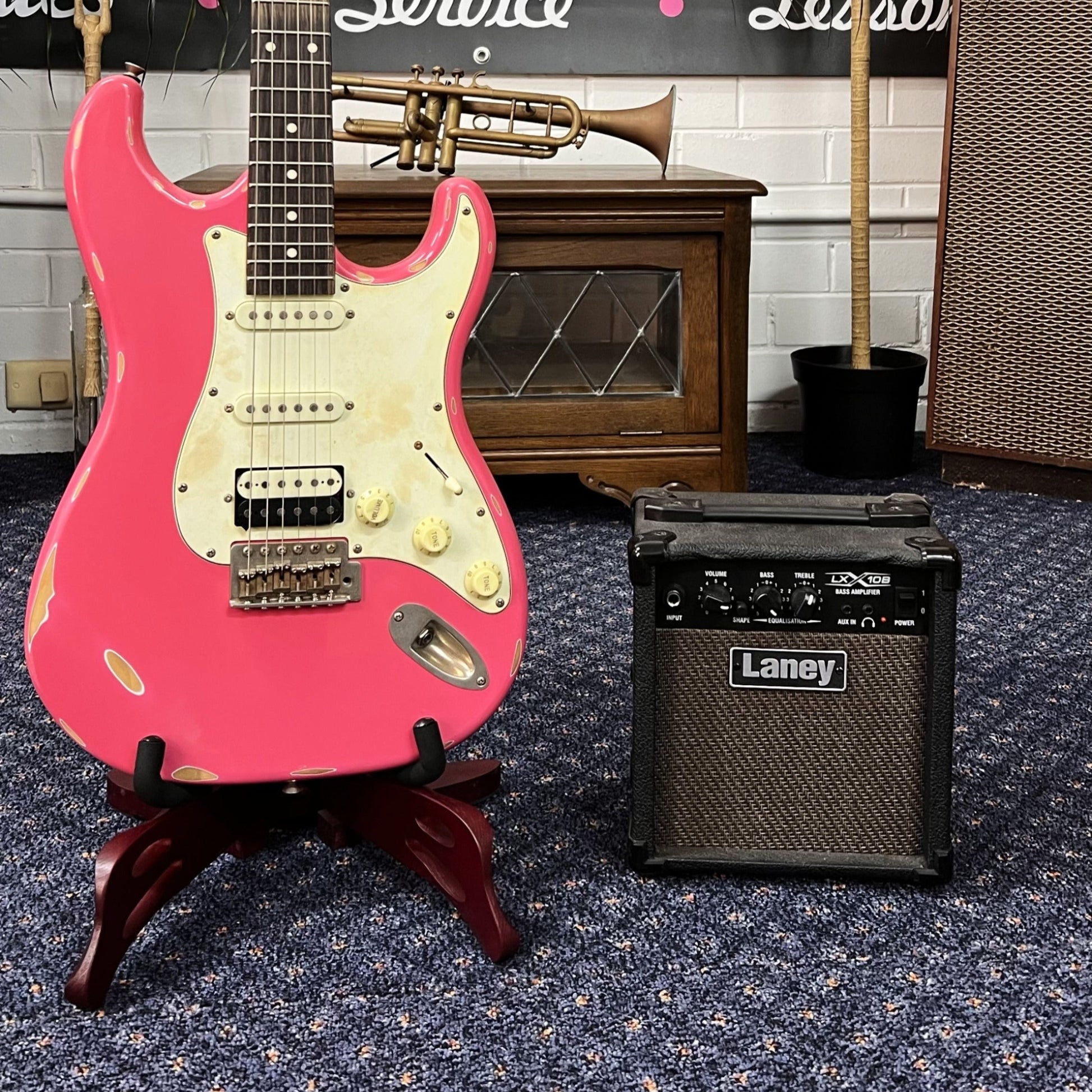 A pink Tokai ST Relic Style electric guitar with a white pickguard, displayed on a stand, next to a Laney LX15 guitar amplifier, all placed on a blue carpeted floor against a backdrop featuring the brand name.